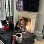 A man sits on a couch beside a fireplace, preparing to clean the chimney
